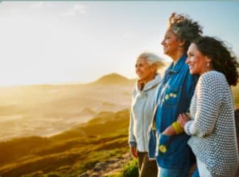 Three women in midlife standing together, looking out over sunlit hills at golden hour